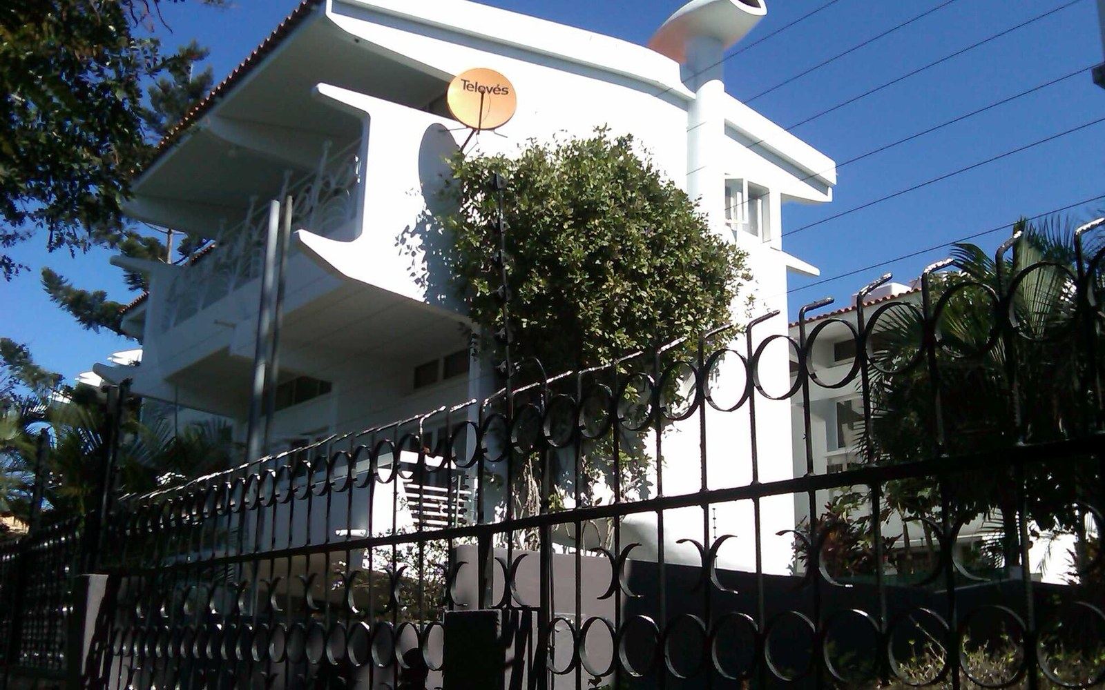 A white modernist villa behind an ironwork fence, deep balconies in hard sun